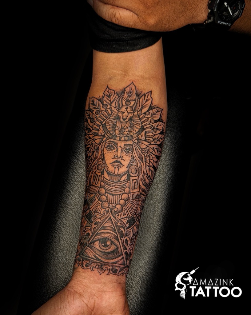 Detailed black and grey forearm tattoo of an Egyptian queen wearing a tribal cat headdress above the Eye of Providence, created by artist Midhun at Amazing Tattoo in Kerala.