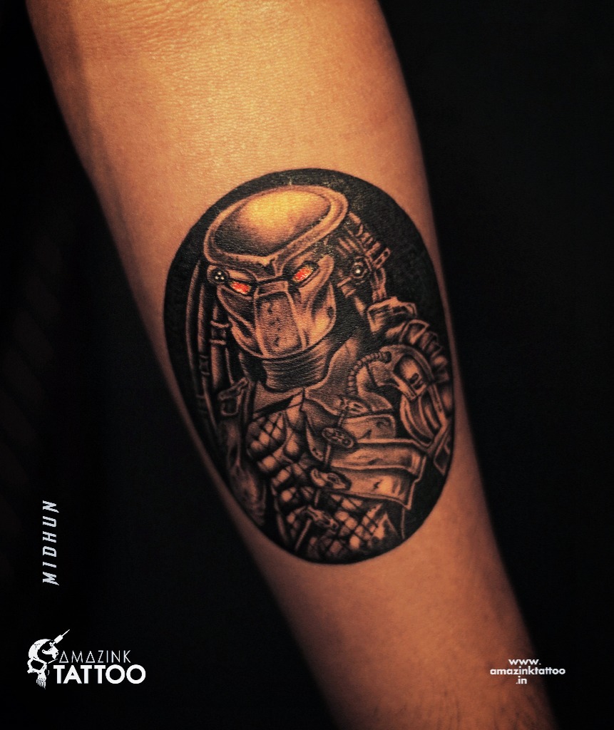 A circular black and grey realism tattoo of the iconic sci-fi Predator character in its biomechanical helmet with glowing red eyes, inked at Amazing Tattoo in Calicut.
