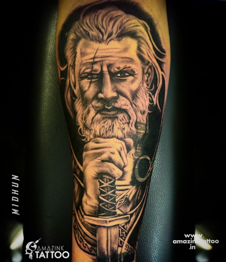 A highly detailed black and grey realism forearm tattoo of a bearded Viking warrior with a facial scar, holding a sword hilt under his chin, custom designed at Amazing Tattoo in Kozhikode.